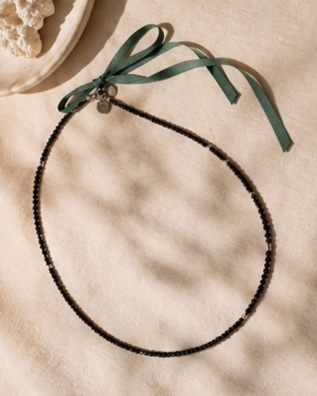 Lange ketting waarin fijne zwarte glaskraaltjes worden afgewisseld met subtiele zilverkleurige metalen accenten en afgewerkt met een donkergroen lint